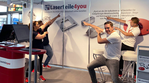 Vier Mitarbeiter am Laserteile4you-Stand zeigen auf die Messewand mit Logo